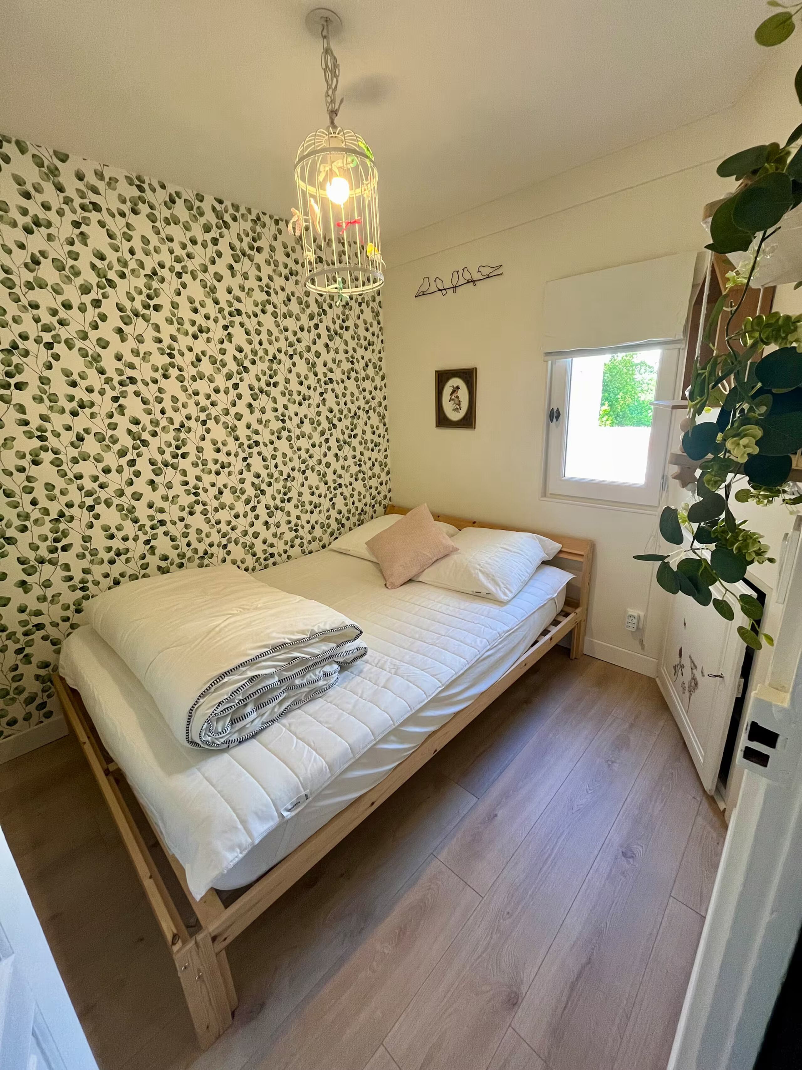 Une chambre avec un lit en bois, un luminaire suspendu, une fenêtre donnant sur un beau paysage et un mur orné d’un motif de feuilles.
