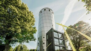 A modern cylindrical building with a flat roof, surrounded by lush greenery and tall grass. - Water tower with Alpine panorama in Chiemgau (Trostberg)