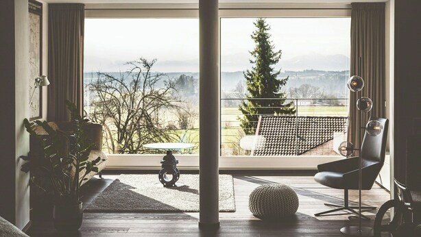 A room with a large window, a black chair, a round ottoman, and a floor lamp. - Water tower with Alpine panorama in Chiemgau (Trostberg)