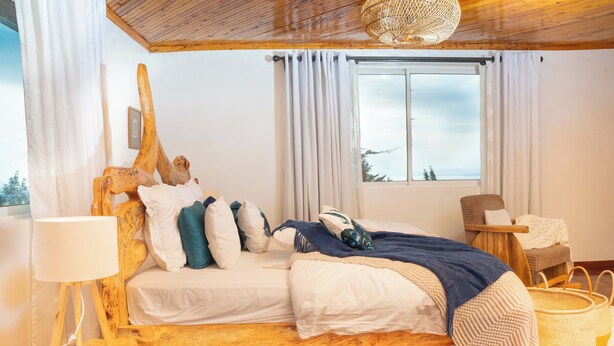 A wooden bed with white bedding, a wooden nightstand, a lamp, a window with curtains, a chair, and a woven basket.