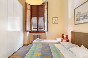 A bedroom with a bed, a radiator, a window with wooden shutters, a bedside table with a lamp, and a framed picture on the wall. - Apartment 'Firenze Famiglie Amici' with Balcony, Wi-Fi and Air Conditioning (Florence)