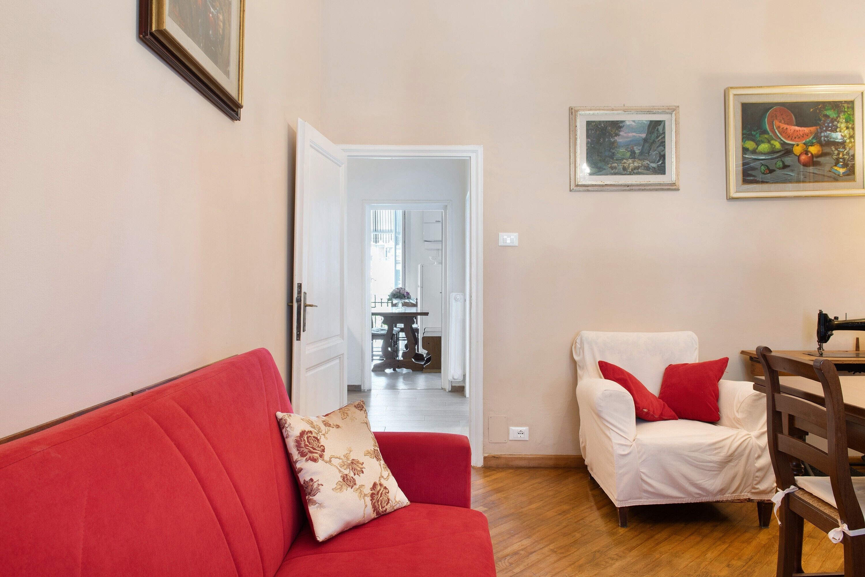 A living room with a red sofa, a white armchair, and a dining table with a sewing machine.