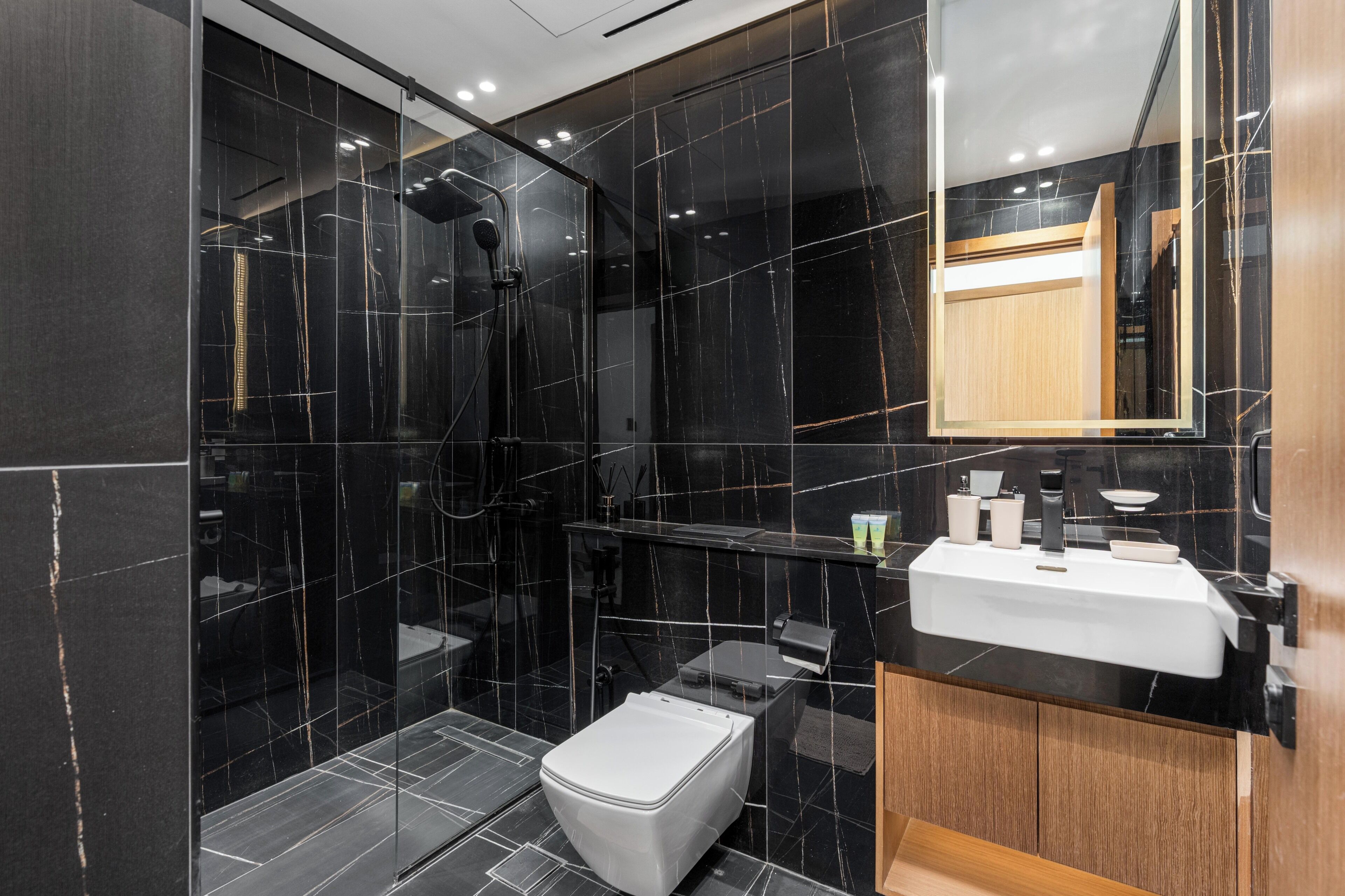 A modern bathroom with a black marble shower, a white toilet, and a wooden vanity with a white sink.