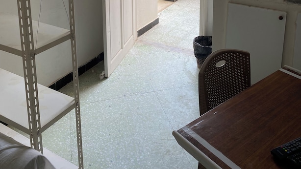 Una habitación con un estante metálico, una mesa de madera, una silla, una puerta blanca que da a un espacio exterior y un lavabo pequeño.