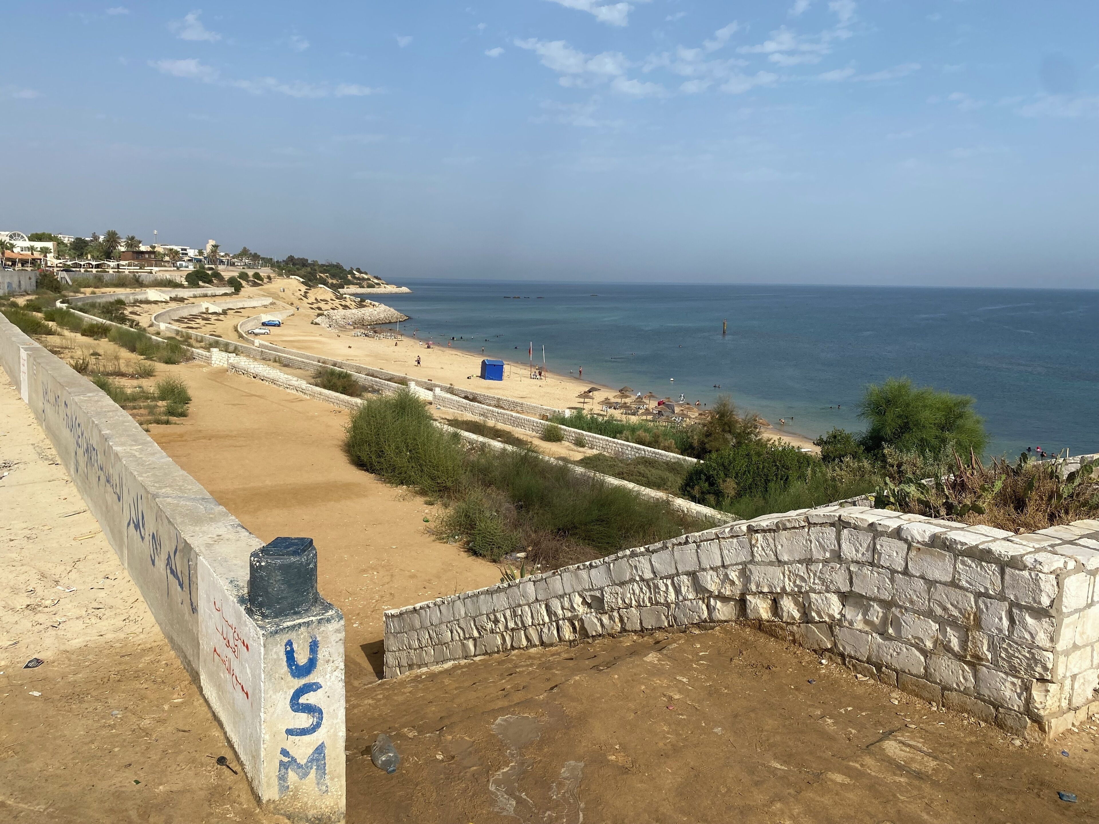 A coastal view with a sandy beach, a stone wall, and a clear sky.