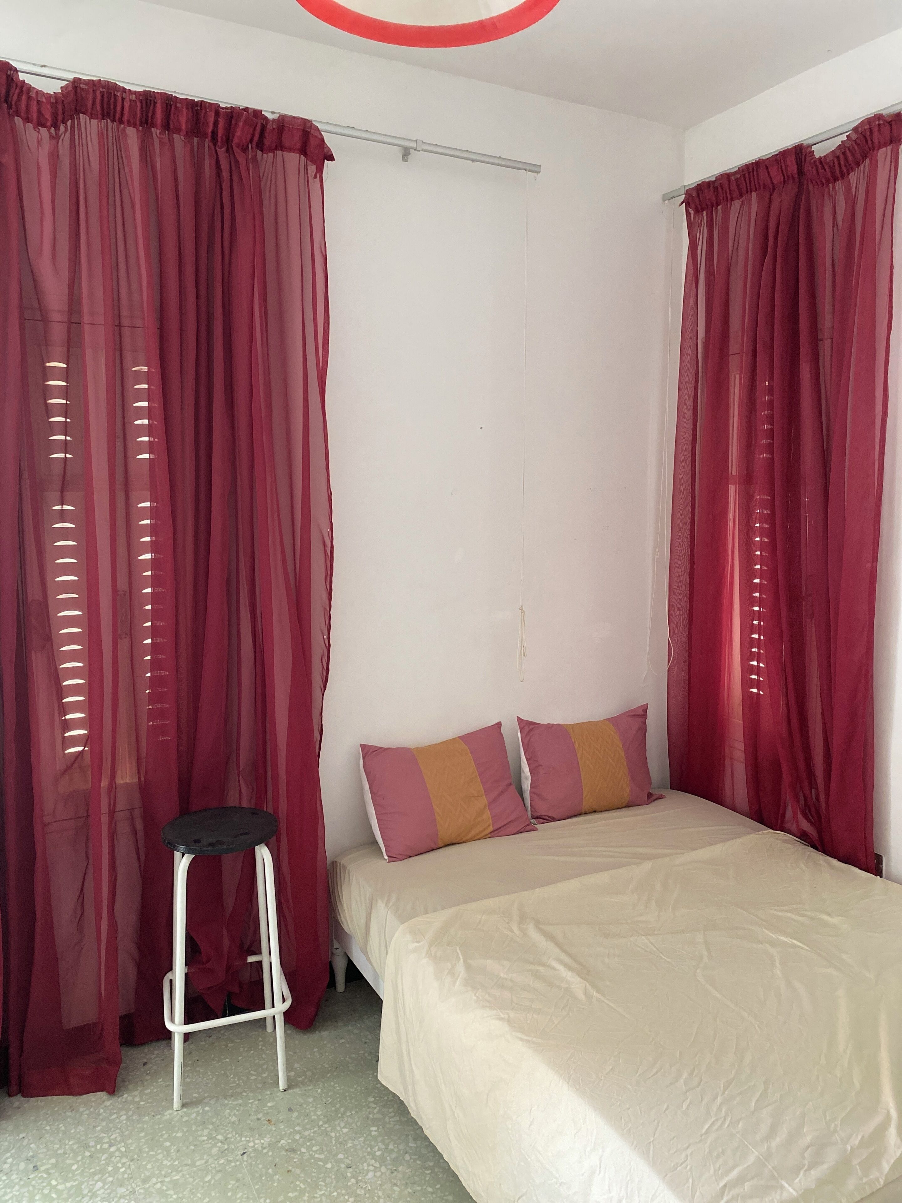 Un dormitorio con una cama, dos almohadas, un taburete y cortinas rojas.