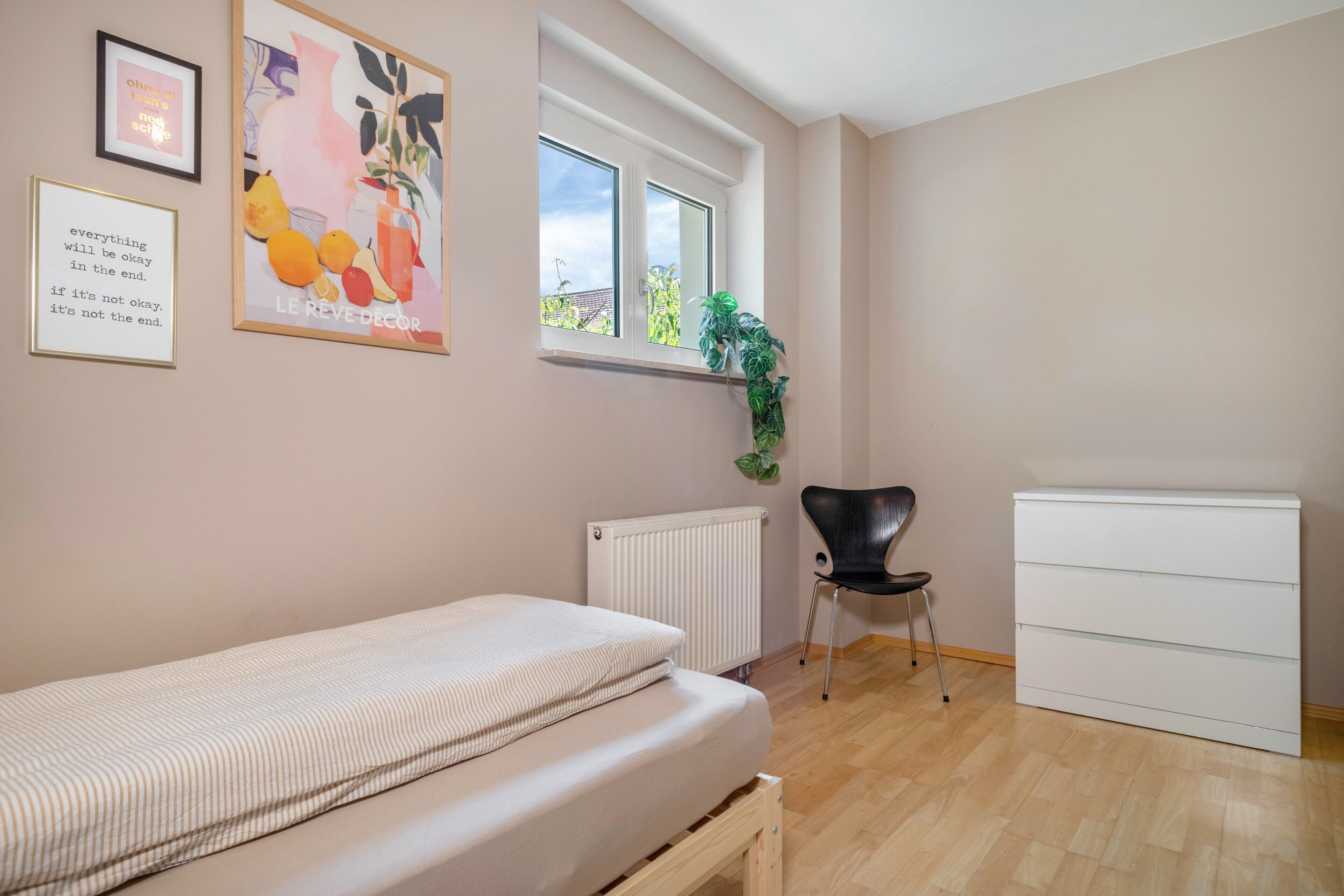 A room with a bed, a radiator, a chair, a white cabinet, and a window with a view of greenery.