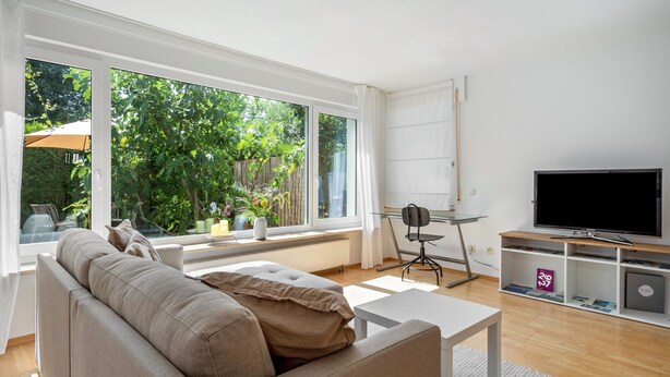 Una sala de estar moderna con un sofá, una mesa de centro, un televisor y un amplio ventanal con vistas a zonas verdes.