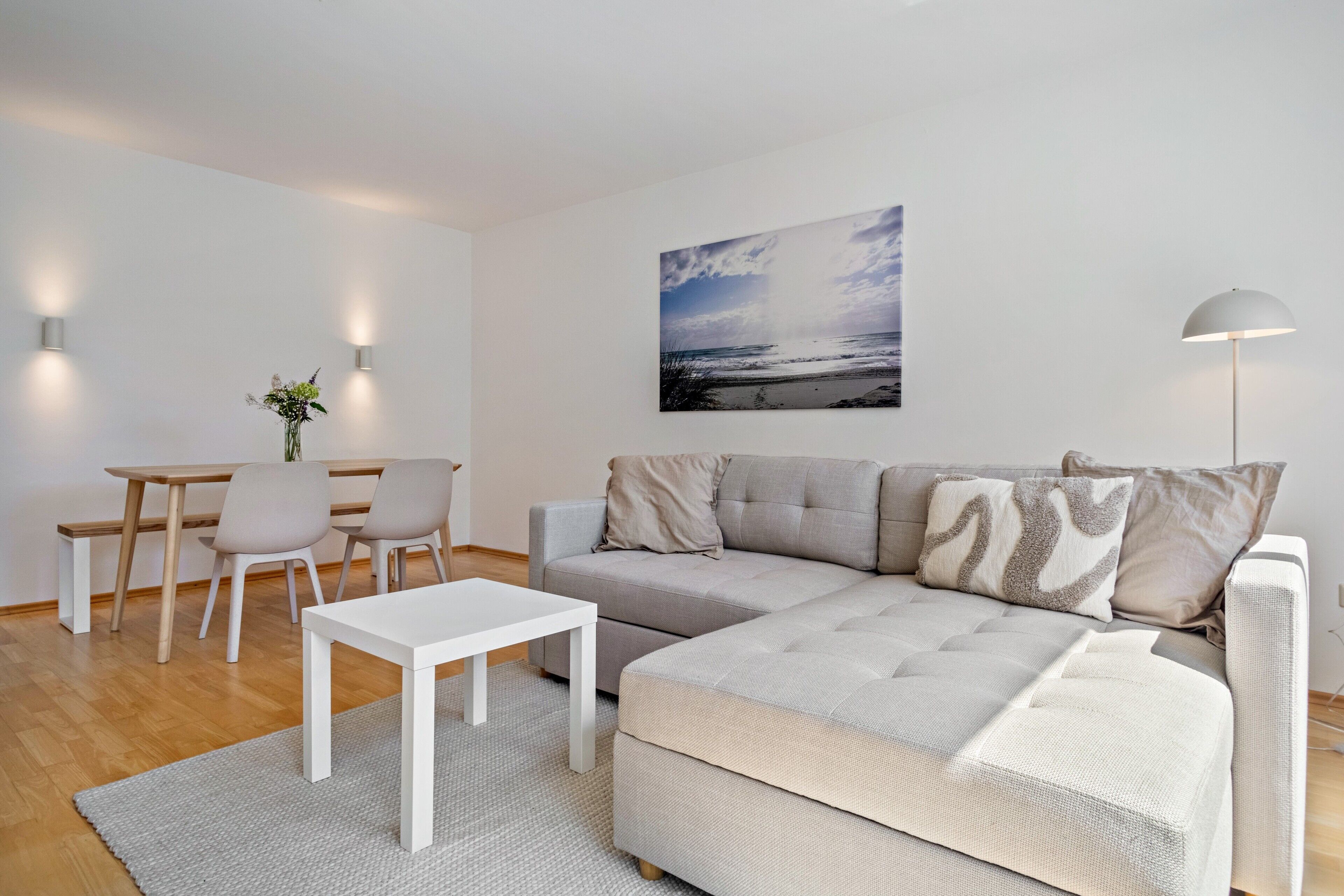 Ett modernt vardagsrum med en grå hörnsoffa, ett vitt soffbord och en matplats med ett litet bord och stolar.