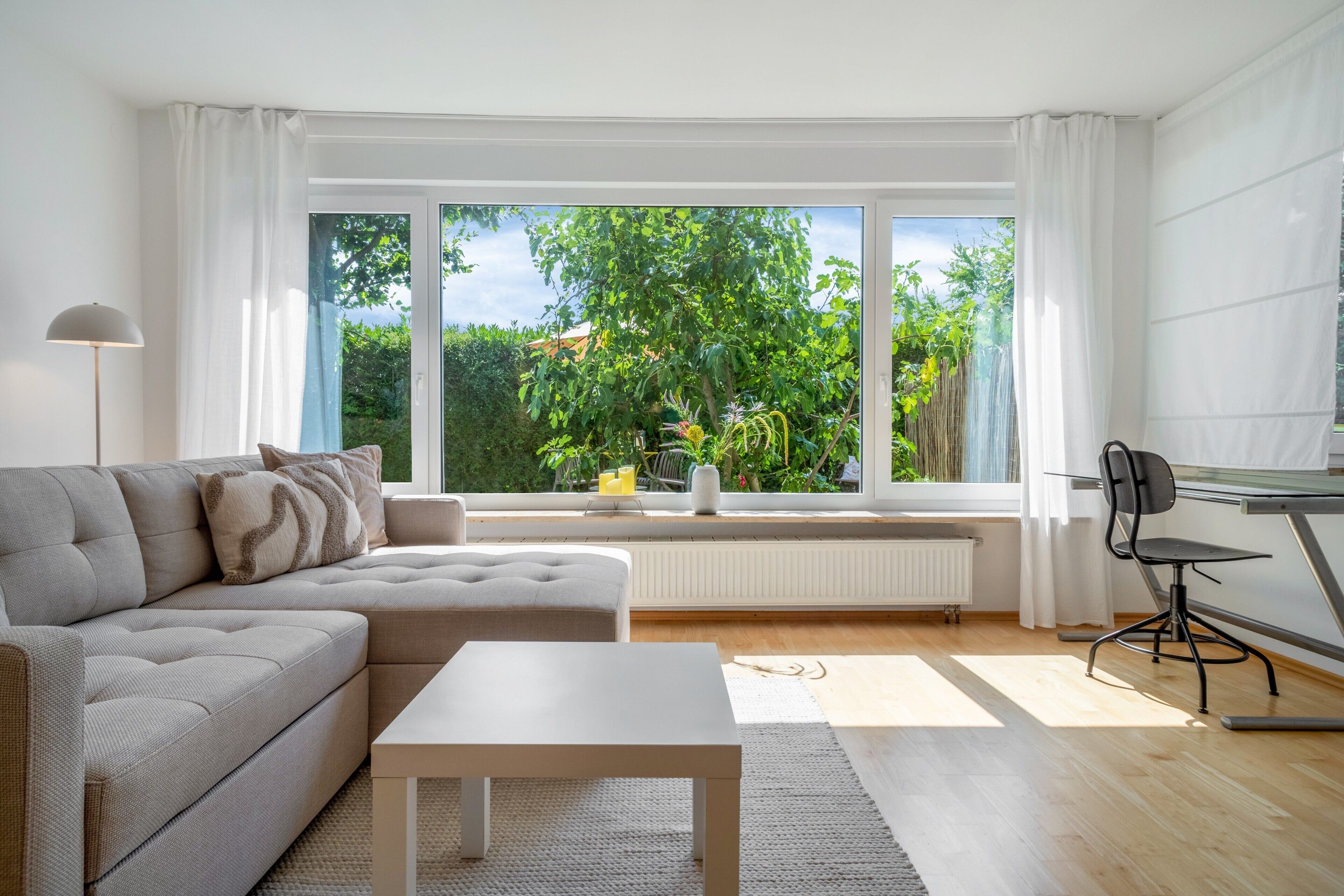 A modern living room with a sofa, a coffee table, and a view of greenery through large windows.