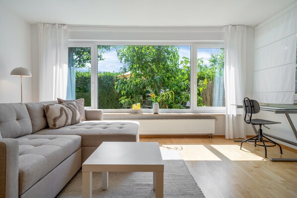 A modern living room with a sofa, a coffee table, and a view of greenery through large windows. - 'Das.møyn' – Your Stay with Mr. Max & Miss Tilda (Friedrichshafen)