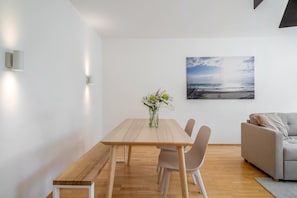 A modern living room with a wooden dining table, two chairs, a grey sofa, and a wall-mounted picture of a beach. - 'Das.møyn' – Your Stay with Mr. Max & Miss Tilda (Friedrichshafen)
