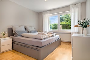 A modern bedroom with a large bed, a bedside table, a dresser, and a window with a view of greenery. - 'Das.møyn' – Your Stay with Mr. Max & Miss Tilda (Friedrichshafen)