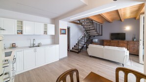 A modern kitchen with white cabinets, a dining area with a wooden table and chairs, a staircase leading to a living area with a white sofa, and a TV on a wooden cabinet. - Casale dei Sogni Entire Private Villa with Terrace, Wi-Fi, and Air Conditioning (Cavaion Veronese)