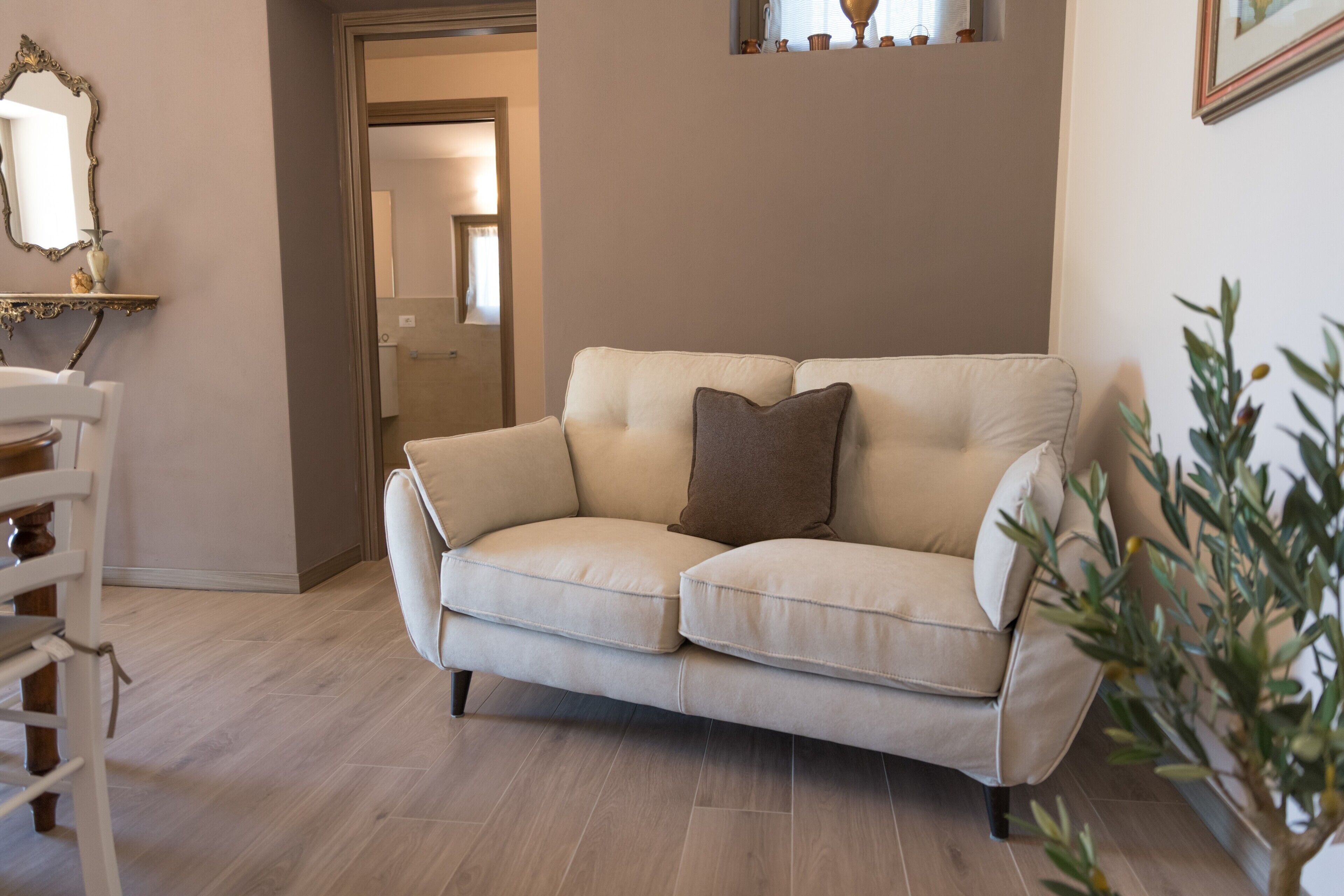 A beige sofa with a brown pillow, a white chair, and a plant in a living room with wooden flooring and a door in the background.