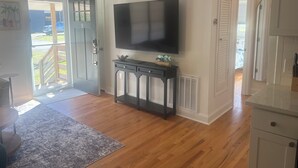 Habitación con televisión de pantalla plana montada en la pared, suelo de madera, puerta que da al exterior y una pequeña consola con objetos decorativos.