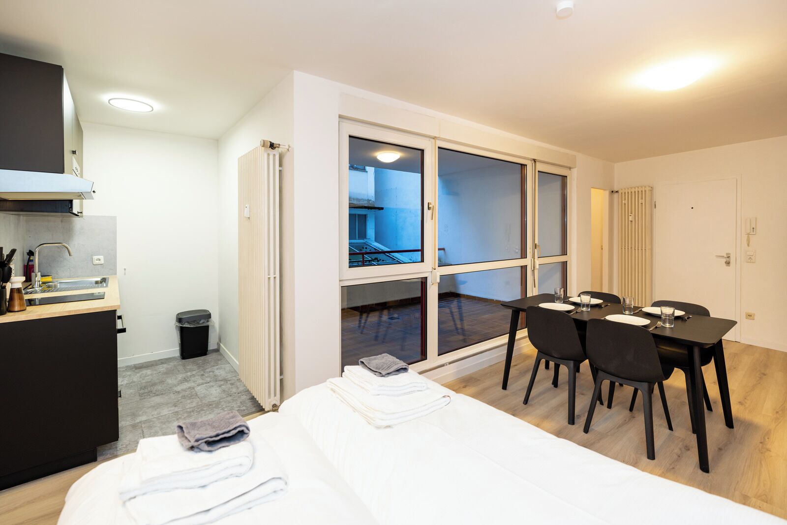 A hotel room with a bed, a dining table with chairs, a kitchen area, and a sliding glass door leading to a balcony.