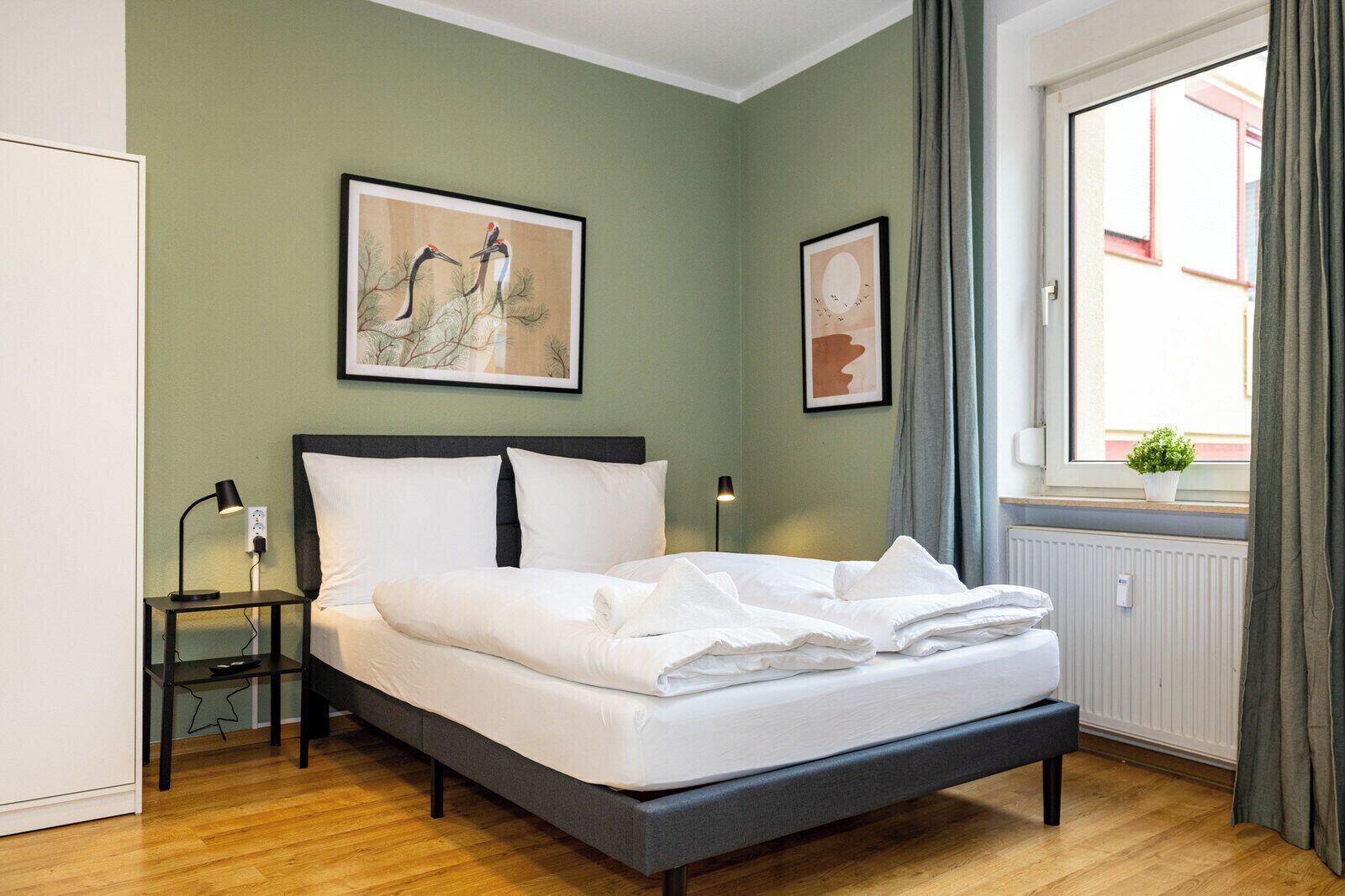 A bedroom with a bed, a bedside table, a lamp, a window with curtains, and a wall-mounted picture.