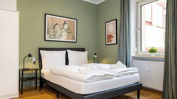 A bedroom with a bed, a bedside table, a lamp, a window with curtains, and a wall-mounted picture.