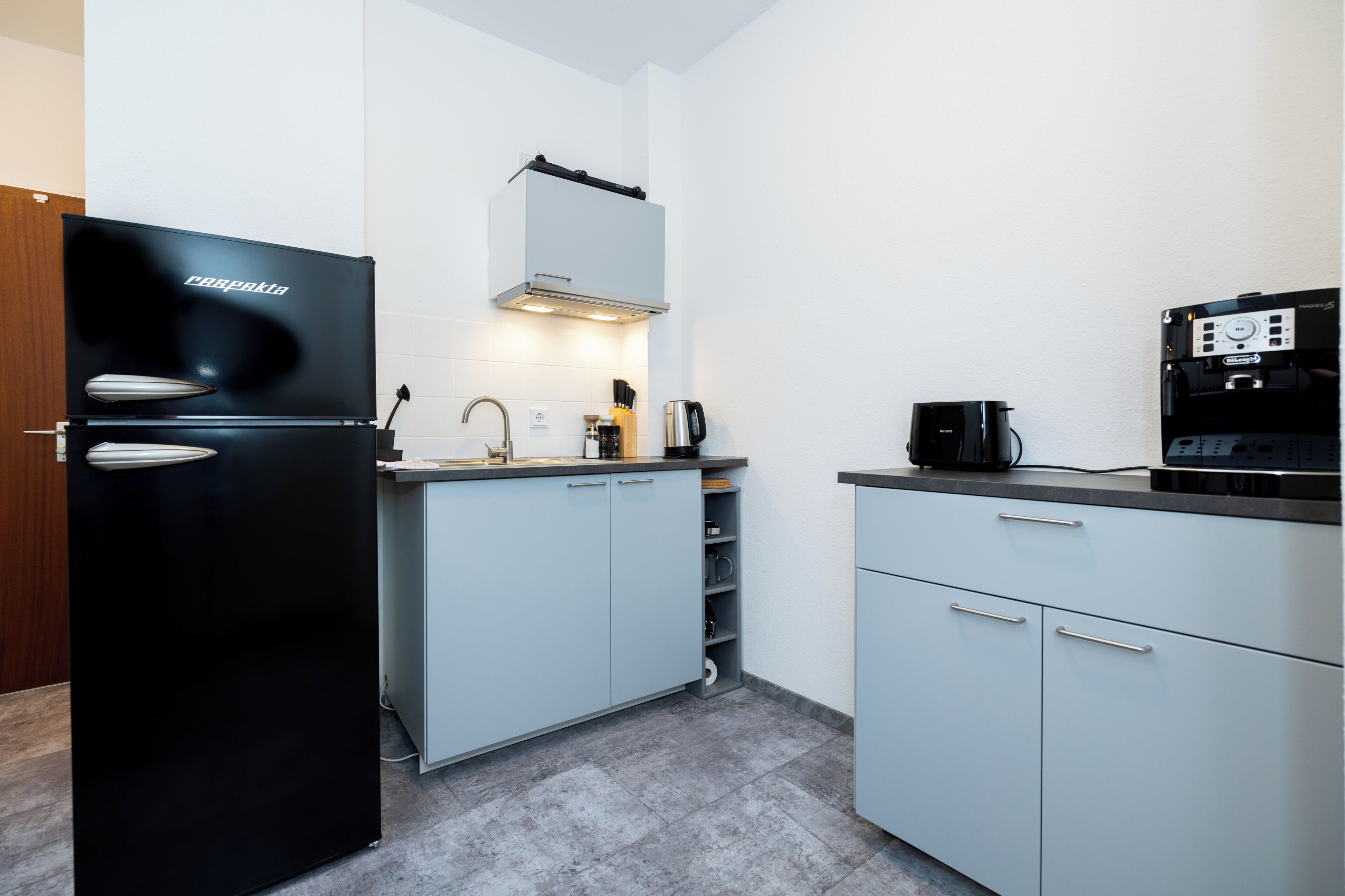 A modern kitchen with a black refrigerator, a coffee machine, and light-colored cabinets.