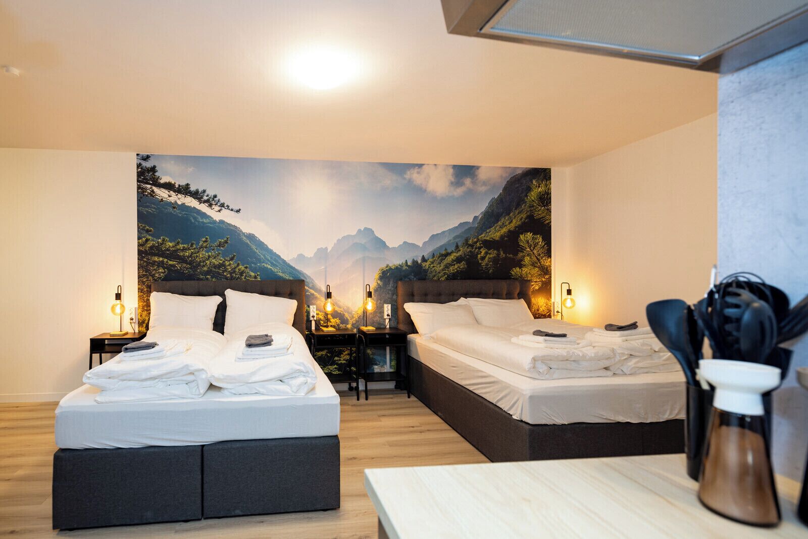 A hotel room with two beds, a wooden floor, a large mountain view mural, and a small table with a coffee pot.