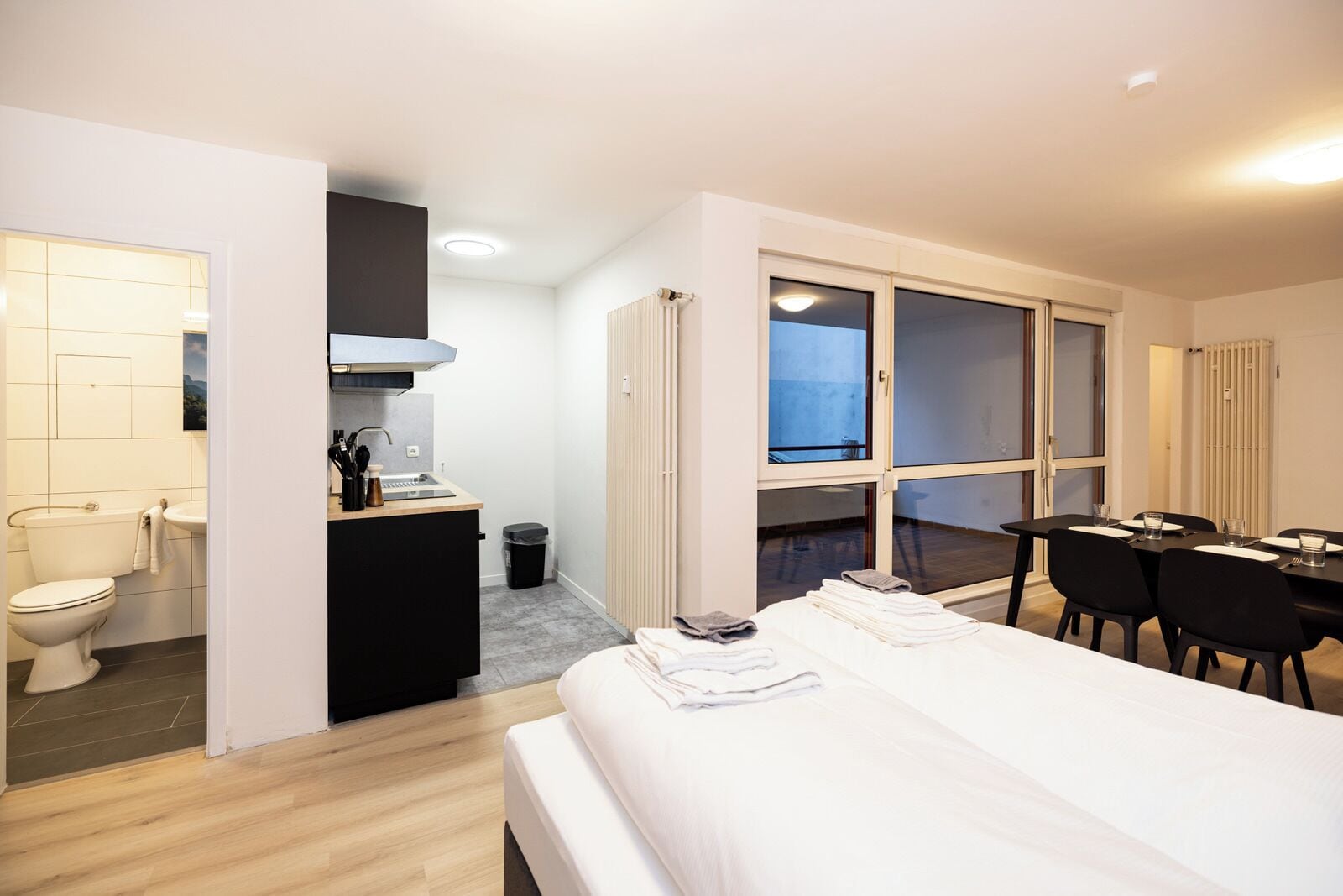 A hotel room with a bed, a small kitchenette, a dining area with a table and chairs, and a large window.