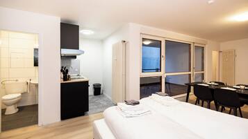 A hotel room with a bed, a small kitchenette, a dining area with a table and chairs, and a large window.