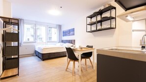 Una habitación de hotel moderna con cama, escritorio y silla, zona de cocina y vistas a la ciudad a través de amplios ventanales.