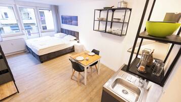 Habitación de hotel pequeña con cama, una mesa de comedor, zona de cocina con lavabo y ventana con vistas a edificios.