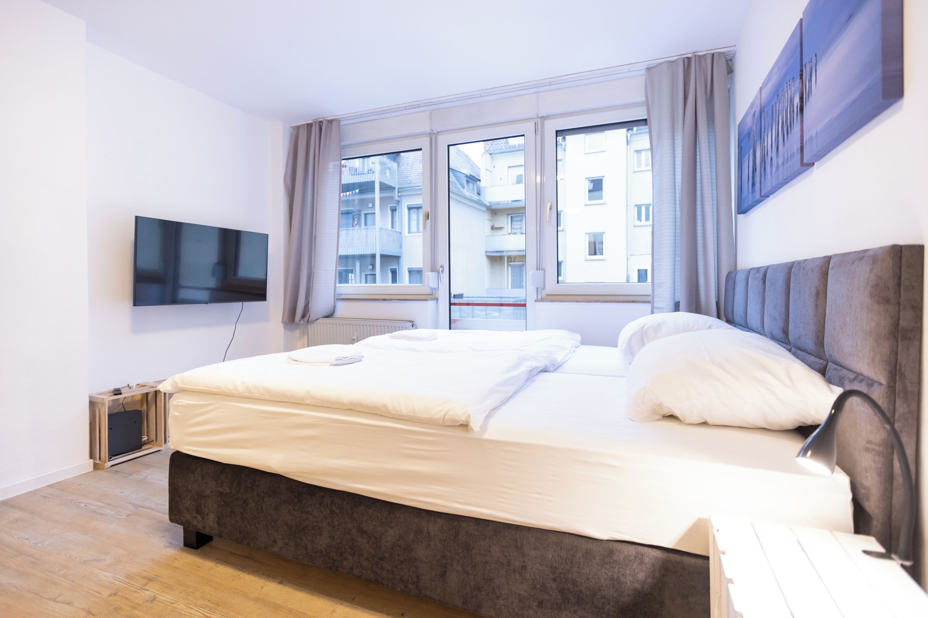 A modern bedroom with a large bed, a flat-screen TV mounted on the wall, and a window with a view of buildings.
