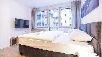A modern bedroom with a large bed, a flat-screen TV mounted on the wall, and a window with a view of buildings.