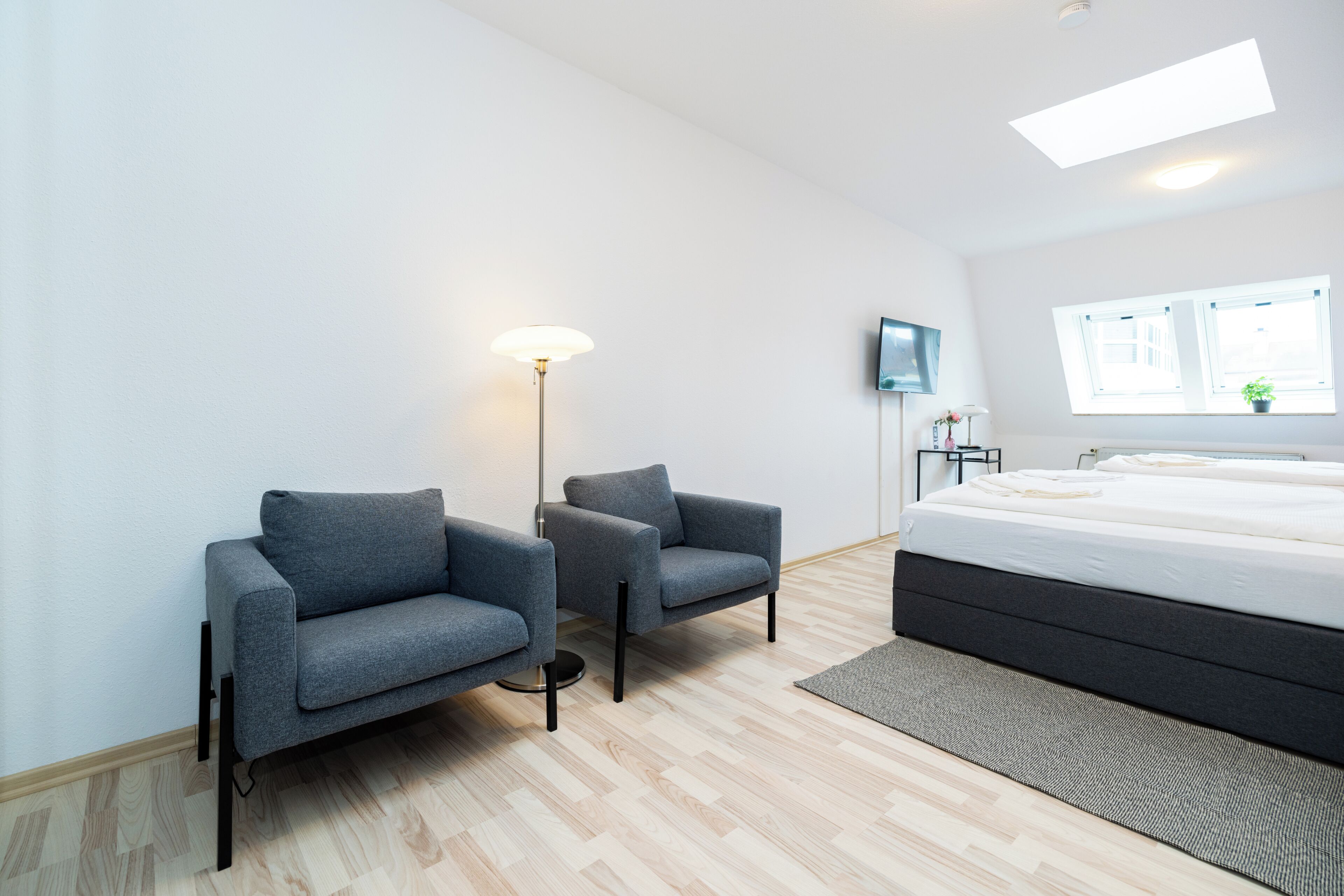A modern bedroom with a bed, two armchairs, a floor lamp, a TV, and a window with a view.