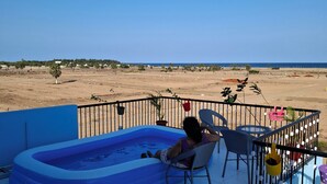 Une personne se détend au bord d’une piscine gonflable bleue, sur un balcon d’où l’on aperçoit une plage et des bâtiments éloignés.