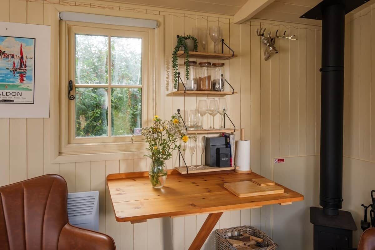 Une cuisine chaleureuse avec une table en bois, un poêle et des étagères où sont disposés de la verrerie et une tête de cerf.