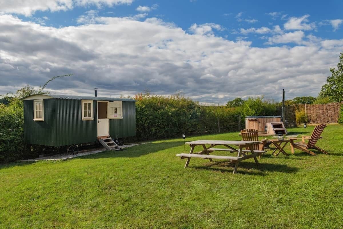 A small green mobile home with a porch and a wooden deck, surrounded by a grassy yard and trees.