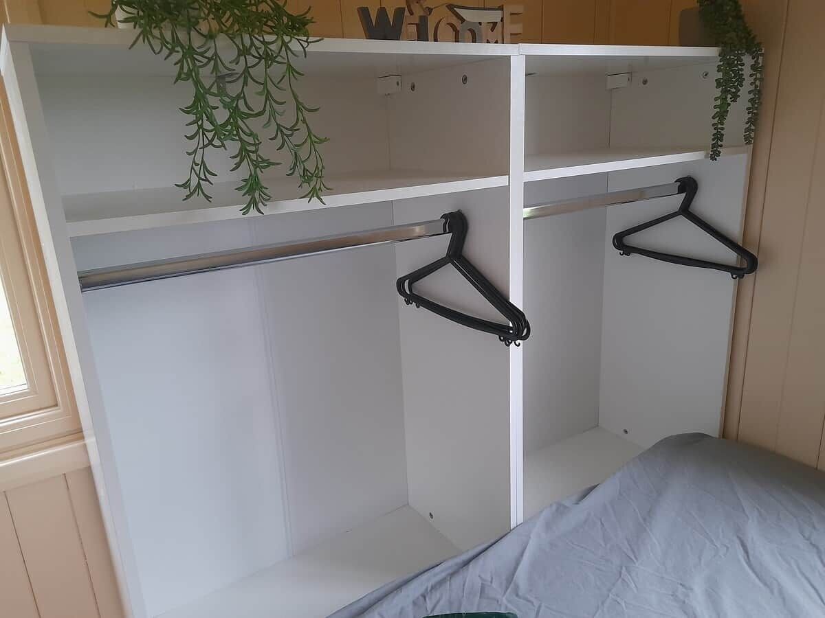 A bedroom with a bed, a wardrobe with two hangers, and a plant on the top shelf.