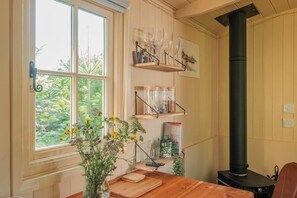 Une pièce avec une table en bois, une fenêtre donnant sur un paysage verdoyant, un poêle noir et des étagères remplies de verrerie et de livres.