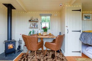A cozy room with a wood-burning stove, two leather chairs, a dining table with breakfast items, and a bed with a blanket. - Hilltop Shepherds hut, hot tub & views of vineyard (Purleigh)