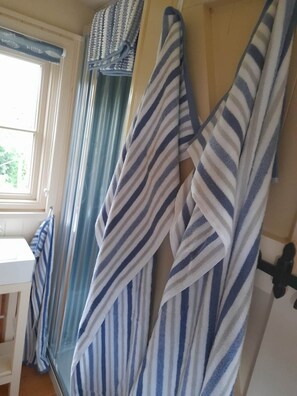 A room with striped towels hanging on a rack, a window with curtains, and a white chair. - Hilltop Shepherds hut, hot tub & views of vineyard (Purleigh)