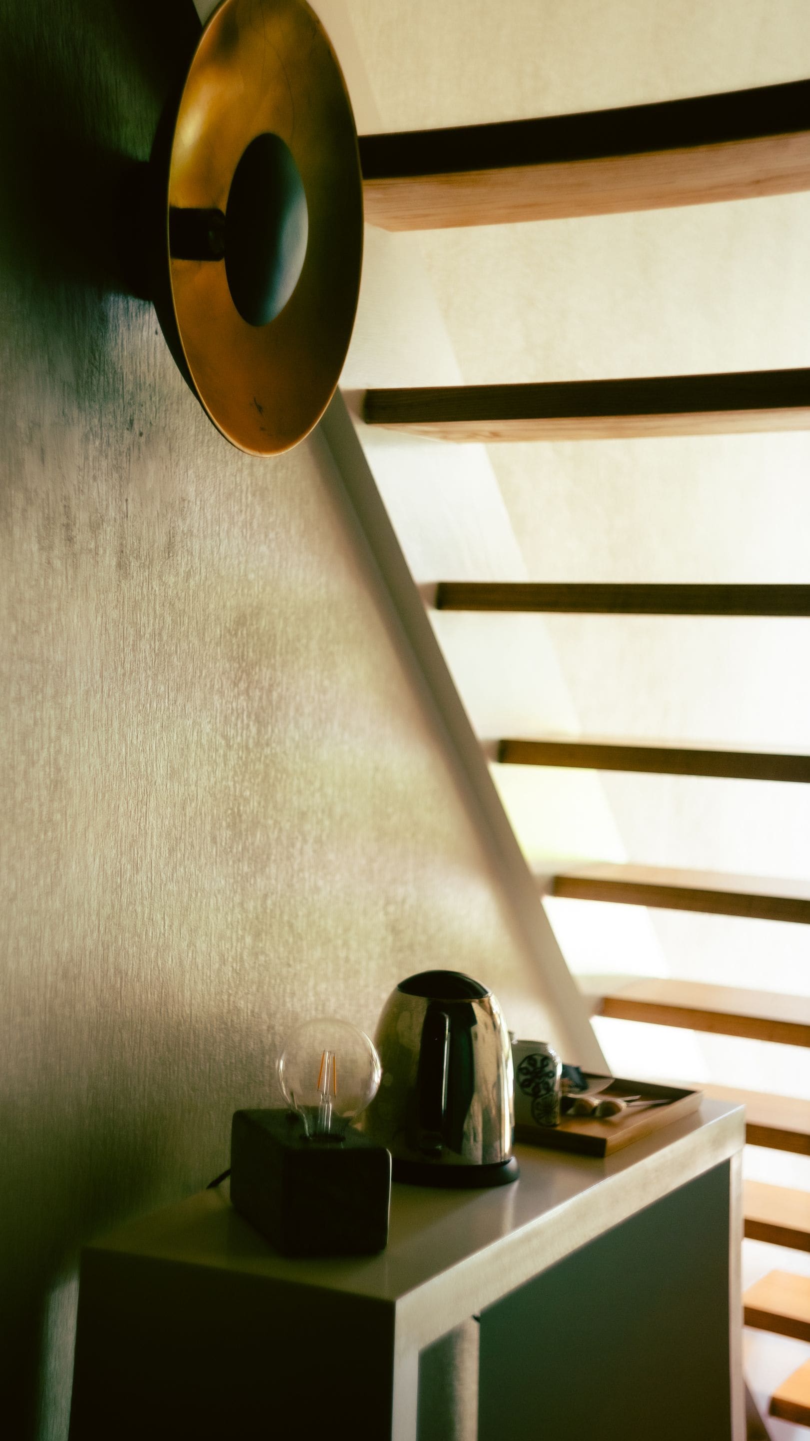 A desk with a lamp, a kettle, and a cup, positioned under a slanted ceiling with wooden beams.