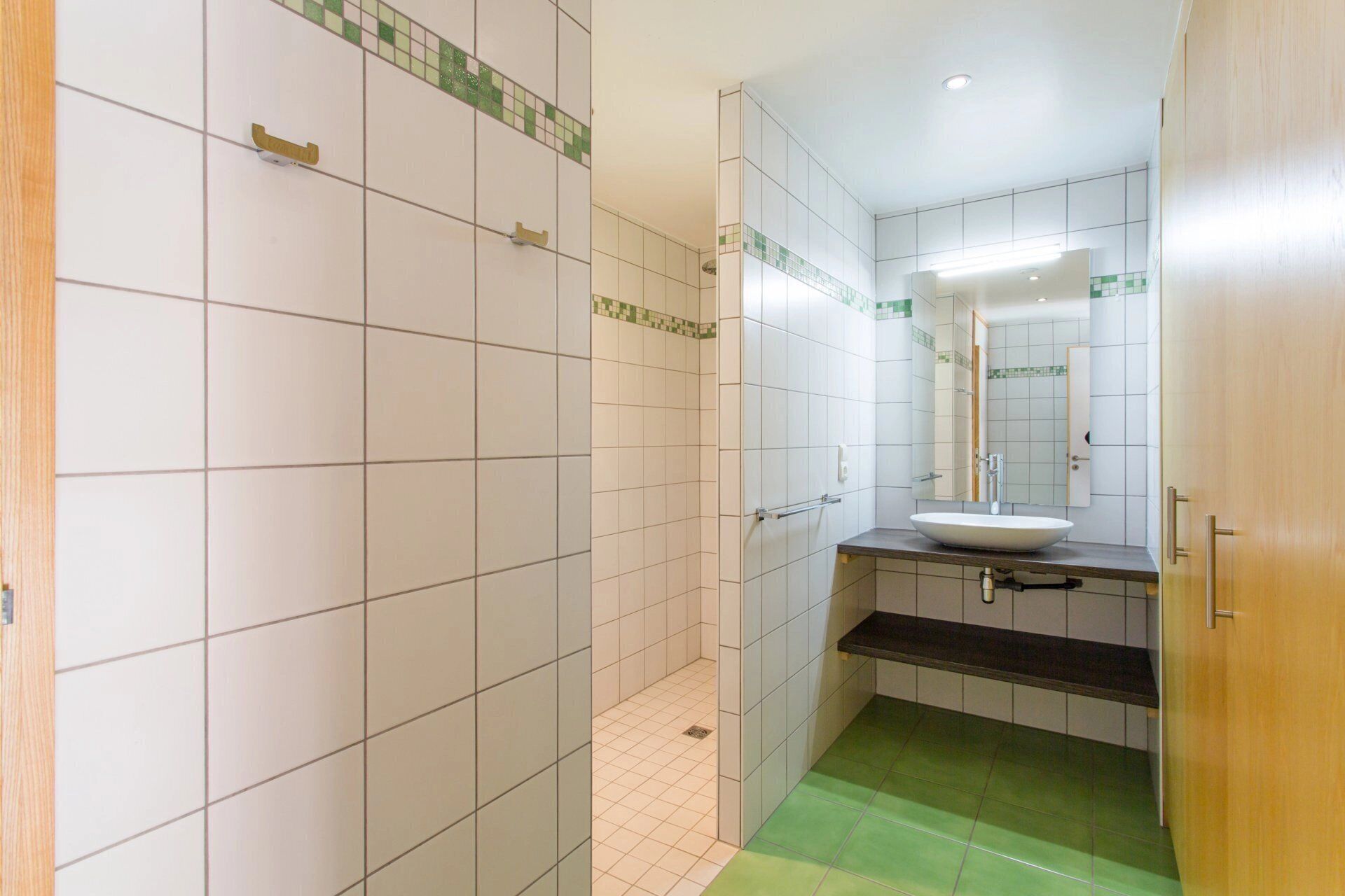 Une salle de bain avec des murs recouverts de carreaux blancs, un sol vert, un lavabo blanc et un miroir.