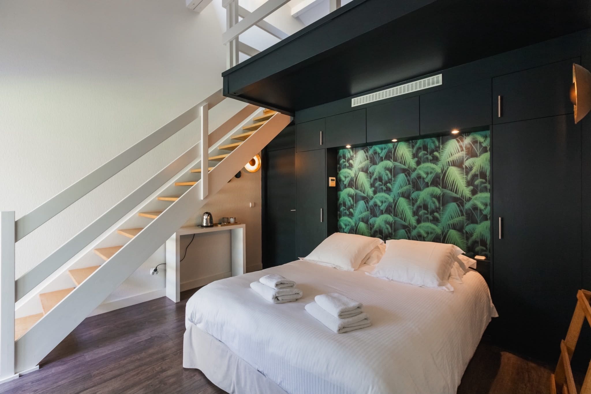 Ein Schlafzimmer mit einem Bett, einem Nachttisch, einem Schreibtisch, einer Treppe und einer Wand mit einem tropischen Blattmuster.