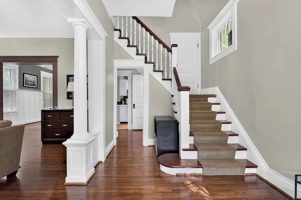 A spacious living room with hardwood flooring, a staircase with a dark wooden handrail, and white walls.