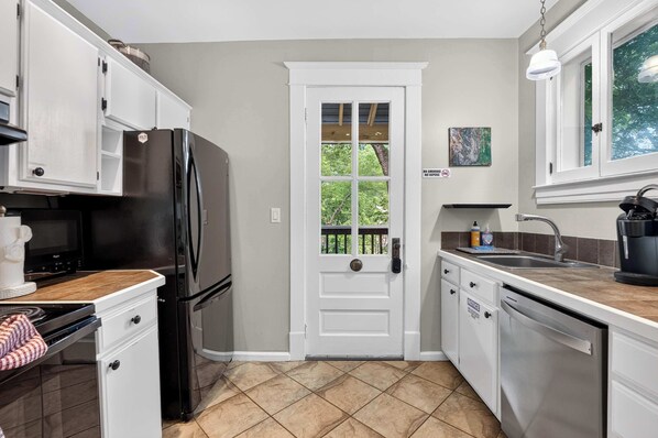 A kitchen with white cabinets, a black refrigerator, a wooden countertop, a sink, and a window with a view of trees.