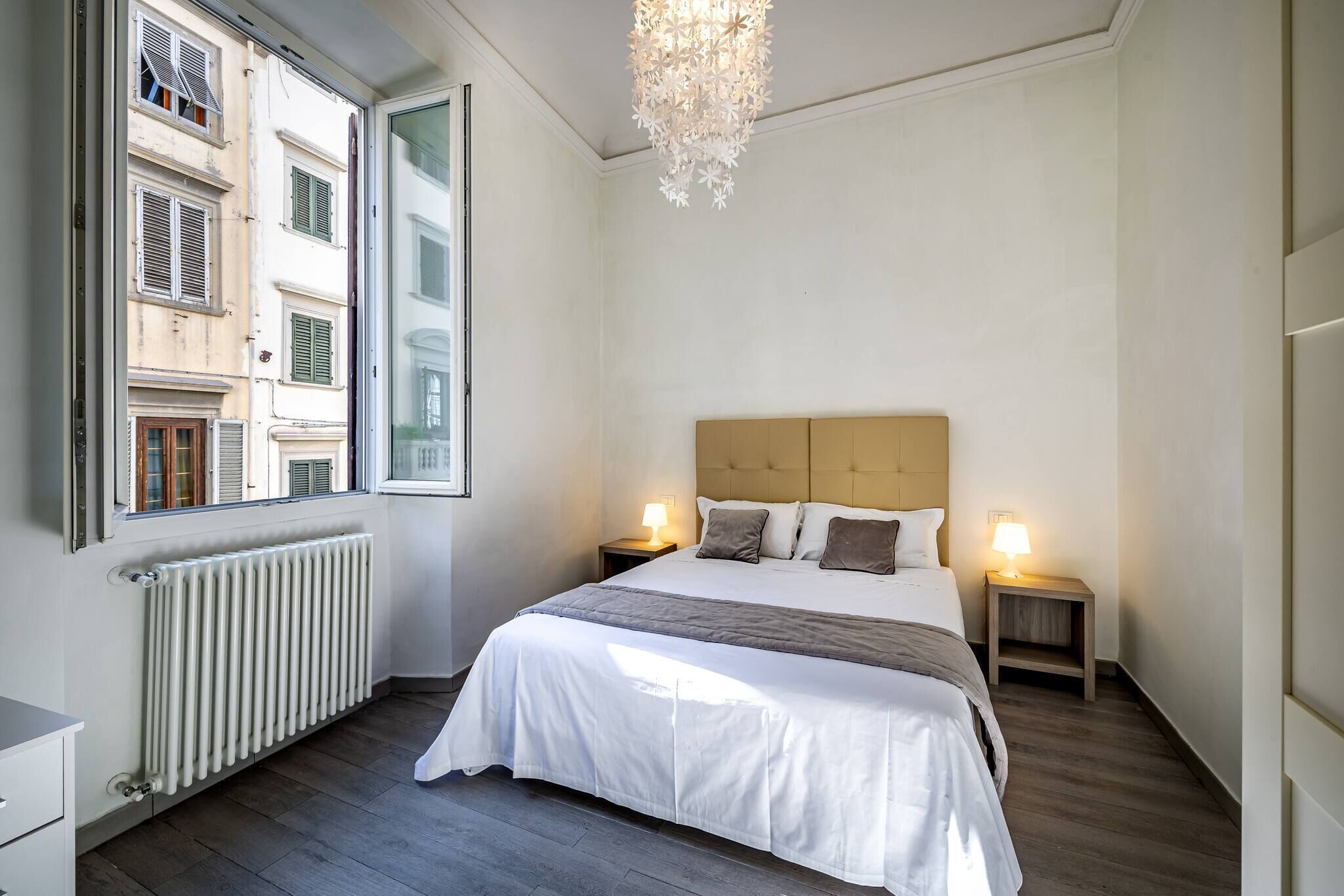 A bedroom with a bed, two bedside tables with lamps, a radiator, and a window with shutters.
