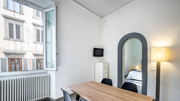 A hotel room with a wooden table, two chairs, a large mirror, a TV on the wall, and a window with a view of buildings. - Proconsolo A (Florence)