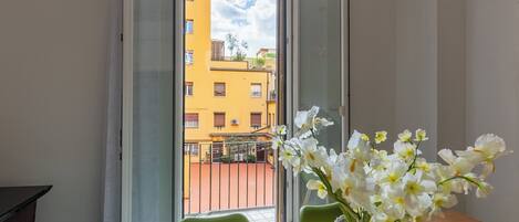 Una habitación con balcón, una mesa con un jarrón de flores y vistas a edificios amarillos.
