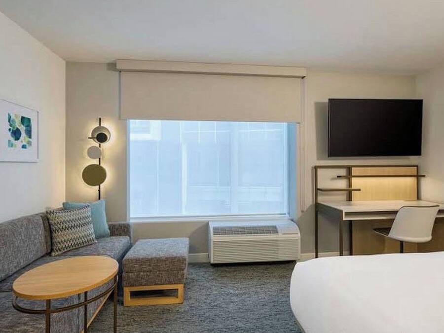 Een hotelkamer met een bank, een klein rond tafeltje, een bed, een bureau met een stoel, een flatscreen televisie en een raam met zonwering.