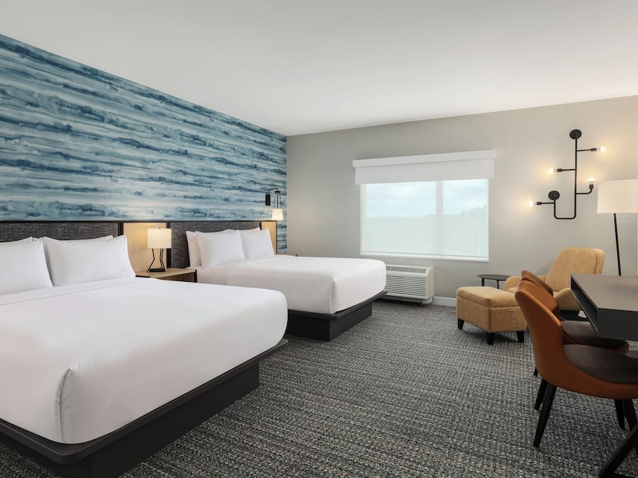 A hotel room with two beds, a desk, a chair, a large window, and a wall with a blue abstract design.