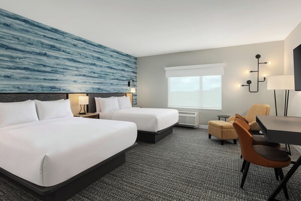 Habitación de hotel con dos camas, un escritorio, una silla, un ventanal grande y una pared con un diseño abstracto en azul.